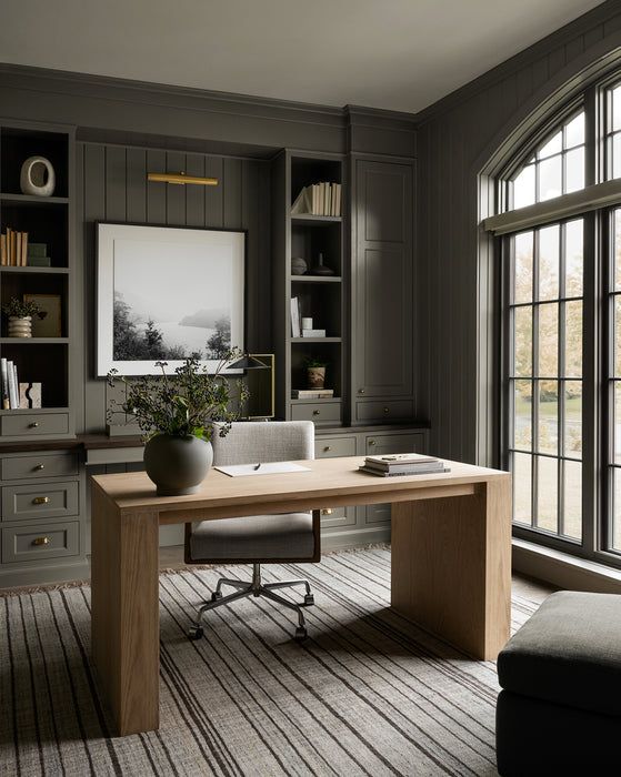 Modern home office with a wooden desk, elegant grey shelves, and large arched window. Comfortable chair and decor accent the minimalistic space.