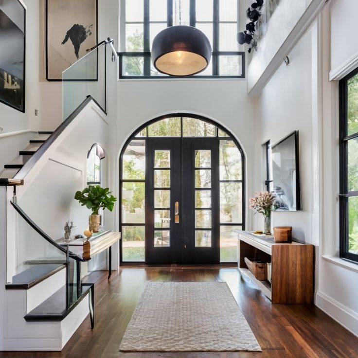 Modern entryway featuring large black doors, wooden flooring, elegant staircase, and stylish decor with natural light streaming through windows.