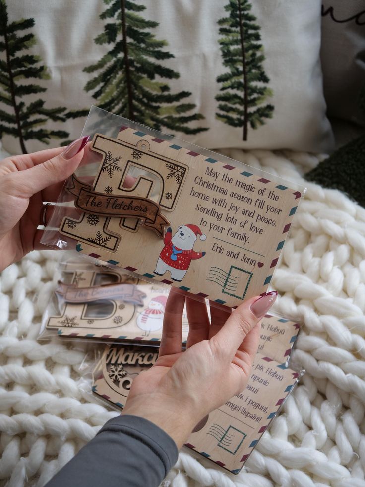 Person holding a personalized wooden Christmas card with festive design and text, in a cozy setting with trees and a knitted blanket.