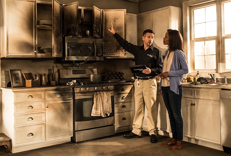 Inspector discussing kitchen fire damage with homeowner, assessing soot-covered cabinets and appliances in bright daylight.