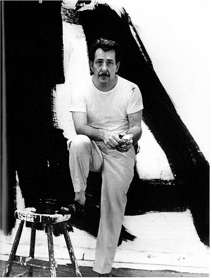 Artist seated on a stool in front of a large abstract black and white painting, wearing a white t-shirt and pants, holding a tool.