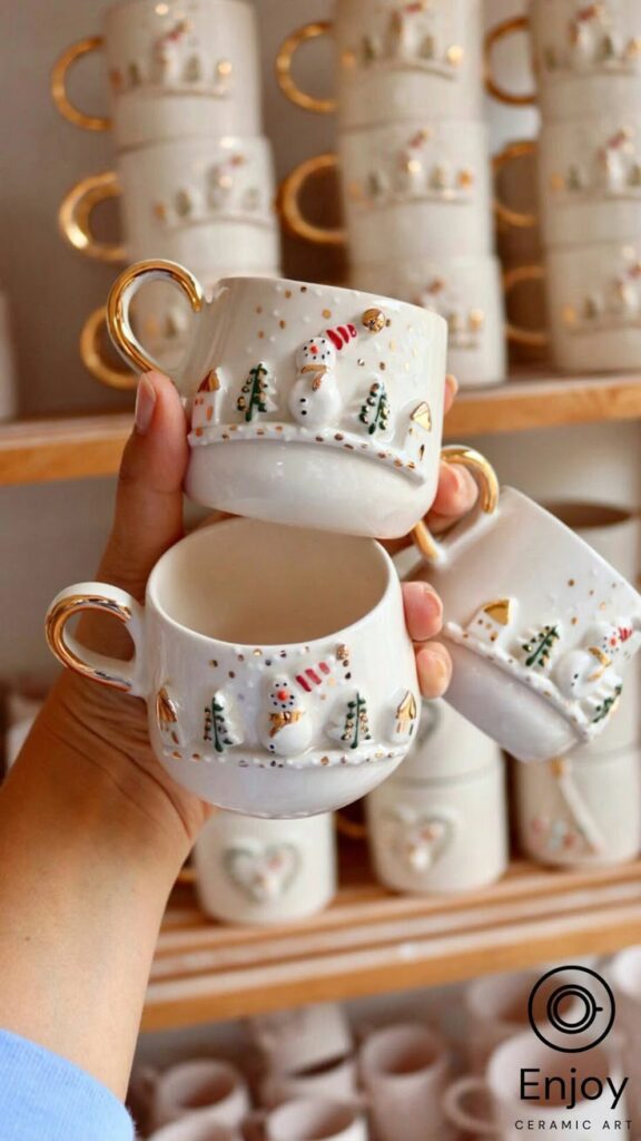 Frosty Charm Snowman Espresso Cup Saucer - 5_4 oz Hand holding two festive ceramic mugs with snowman and tree design, featuring gold accents, against a backdrop of stacked mugs. | Sky Rye Design Hand holding two festive ceramic mugs with snowman and tree design, featuring gold accents, against a backdrop of stacked mugs.