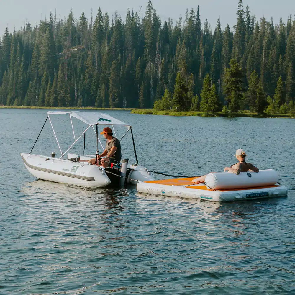 Two people relax on inflatable boats in a serene lake surrounded by lush pine trees under a clear blue sky.