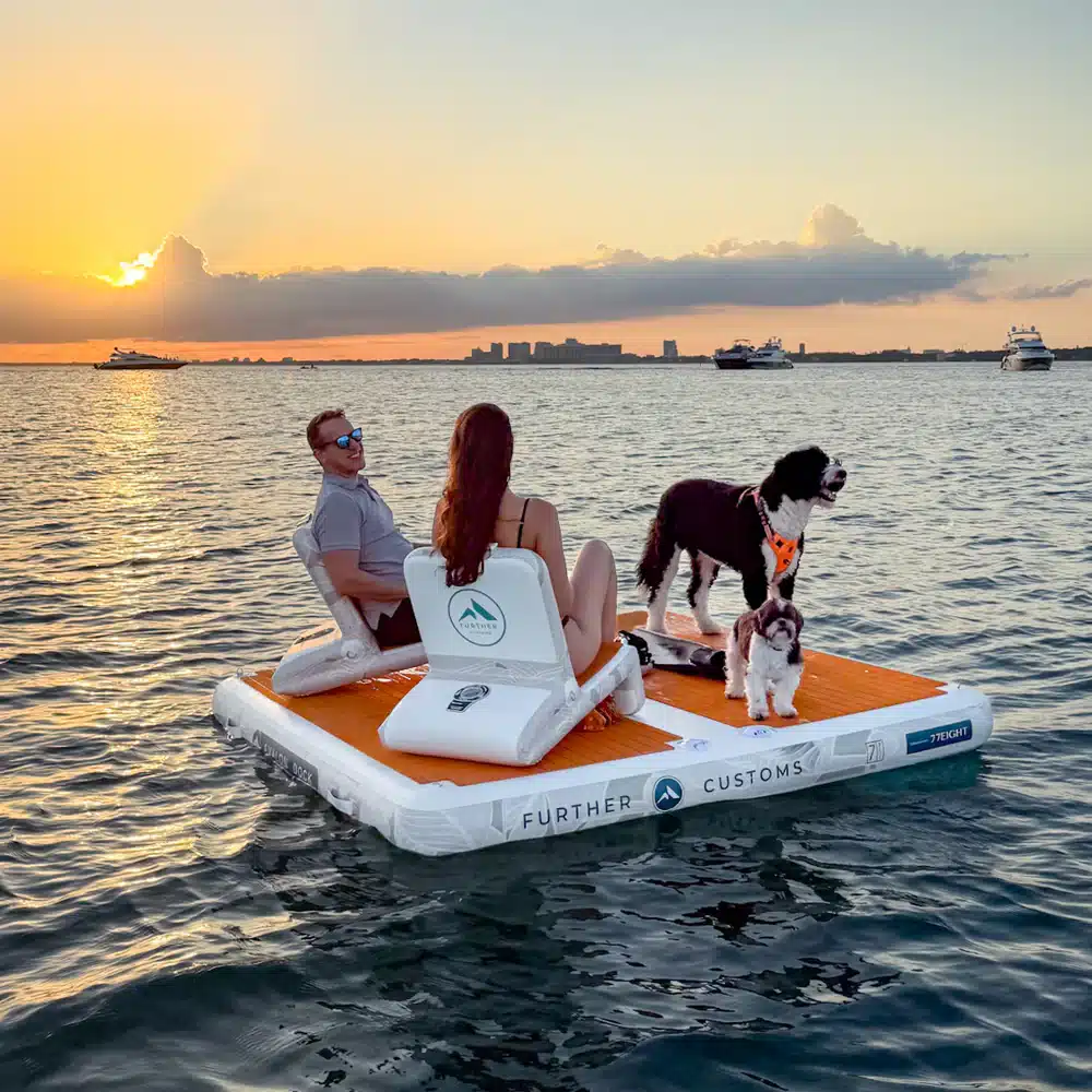 Couple and dogs relaxing on a floating platform during sunset on the ocean, with yachts and city skyline in the distance.