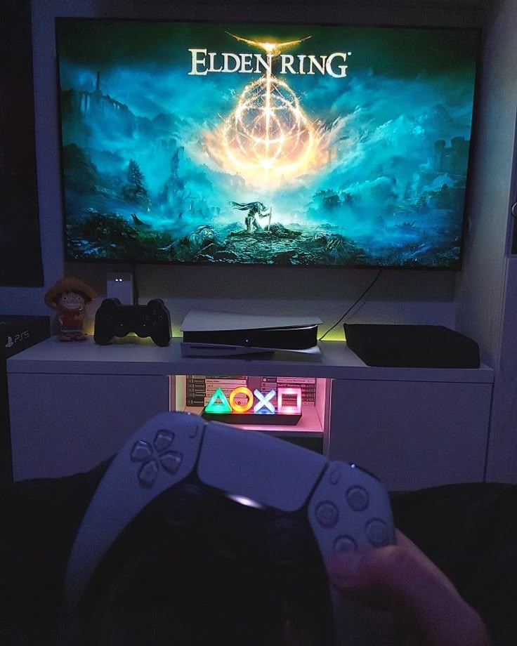 Gamer holding PlayStation controller playing Elden Ring on PS5, vibrant game screen and illuminated PS symbols in cozy setup.