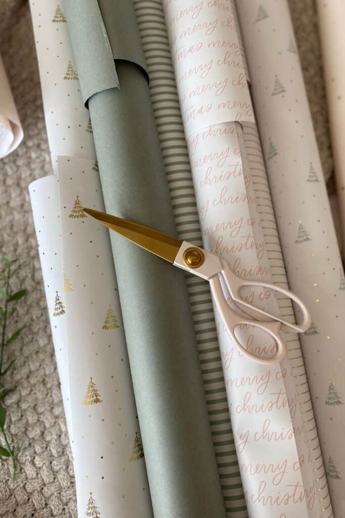 Gift Wrapping Techniques _ Mornington Lane Assorted Christmas wrapping paper rolls with a gold and white scissors on a textured surface, ready for gift wrapping. | Sky Rye Design Assorted Christmas wrapping paper rolls with a gold and white scissors on a textured surface, ready for gift wrapping.