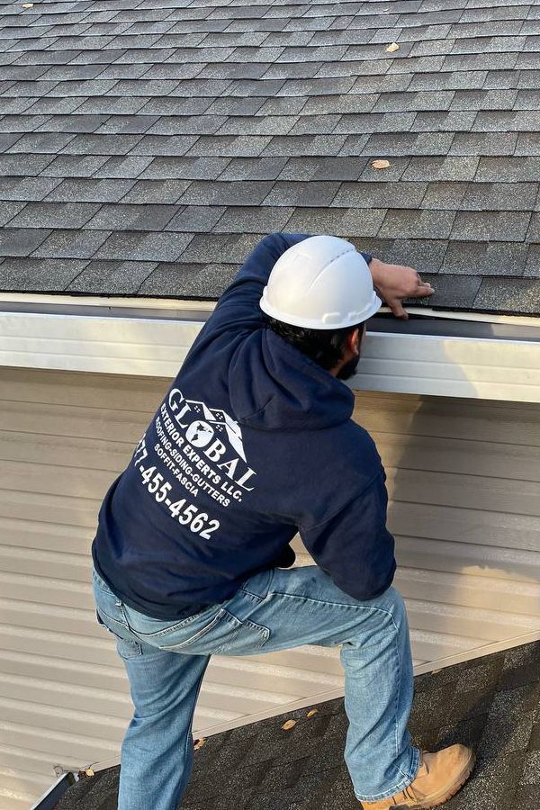 Global Exterior Experts _ Roof Inspection Worker inspecting roof shingles while wearing a hard hat and blue hoodie, ensuring safety and maintenance. | Sky Rye Design Worker inspecting roof shingles while wearing a hard hat and blue hoodie, ensuring safety and maintenance.