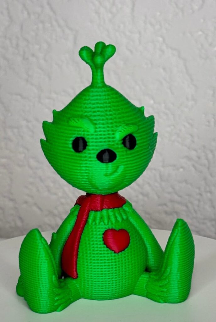 Green 3D-printed character with red scarf and heart sitting against white textured wall.