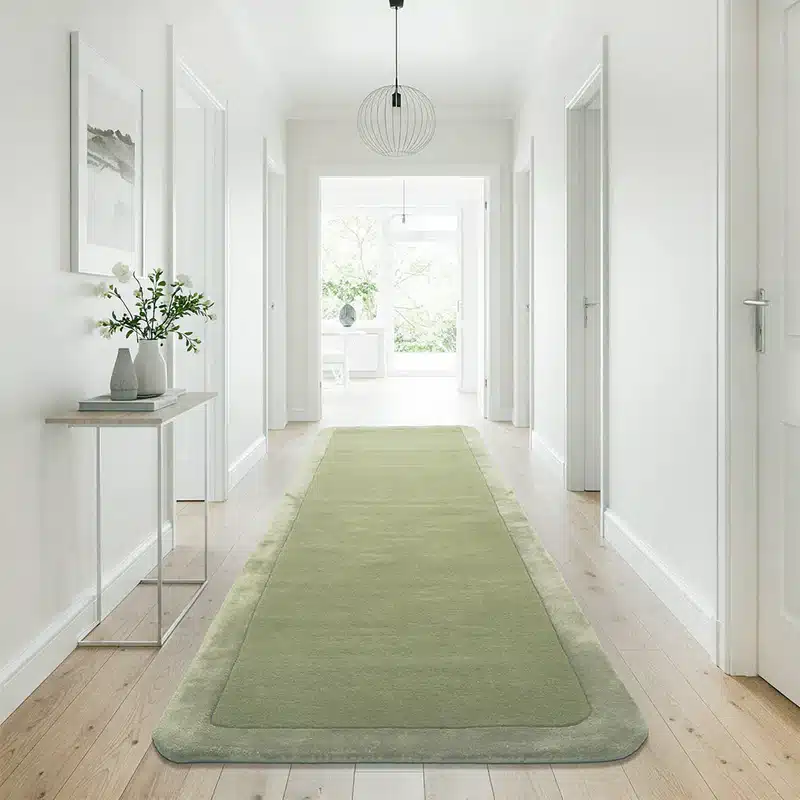 Modern hallway with light wood flooring, green runner rug, minimalist decor, and natural light through large windows.