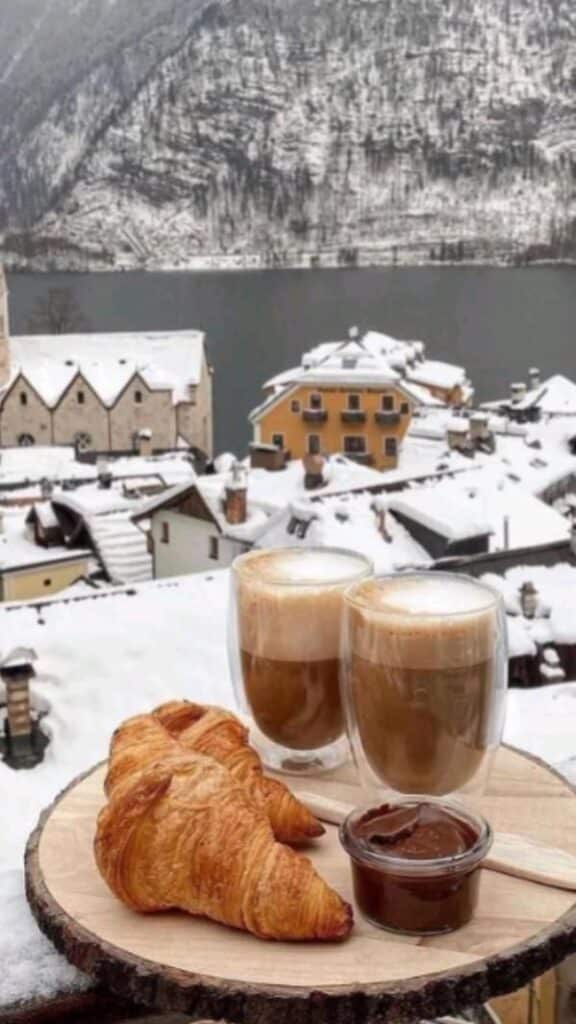 Snowy village scene with croissants, cappuccinos, and chocolate spread, overlooking a lake and snow-covered rooftops in winter.