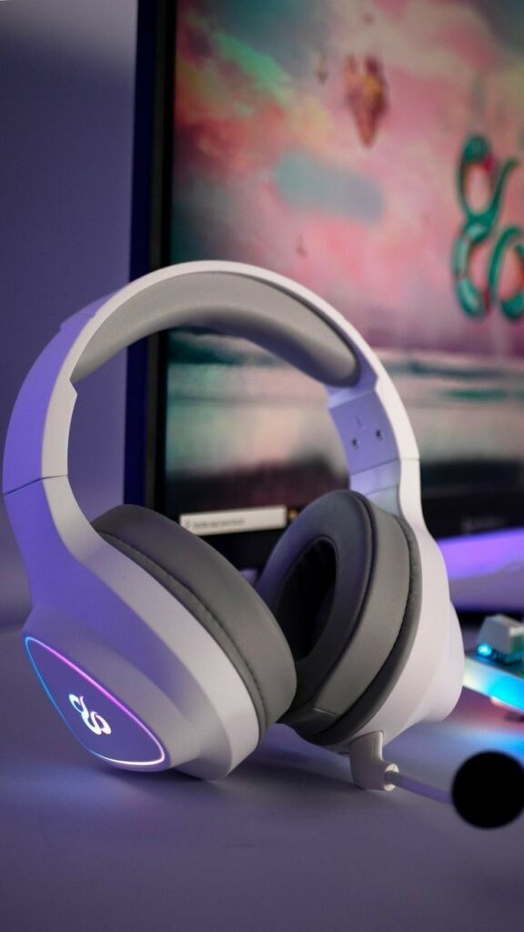 White gaming headset with LED lighting on a desk, in front of a colorful monitor display, creating an immersive gaming setup atmosphere.
