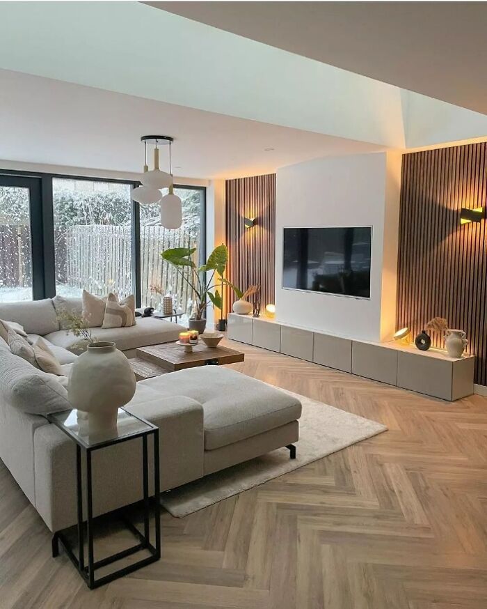 Modern living room with cozy beige sofa, wooden accents, large TV, and natural light from floor-to-ceiling windows.
