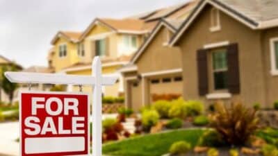 For sale sign in front of suburban house, indicating property available for purchase in a residential neighborhood.
