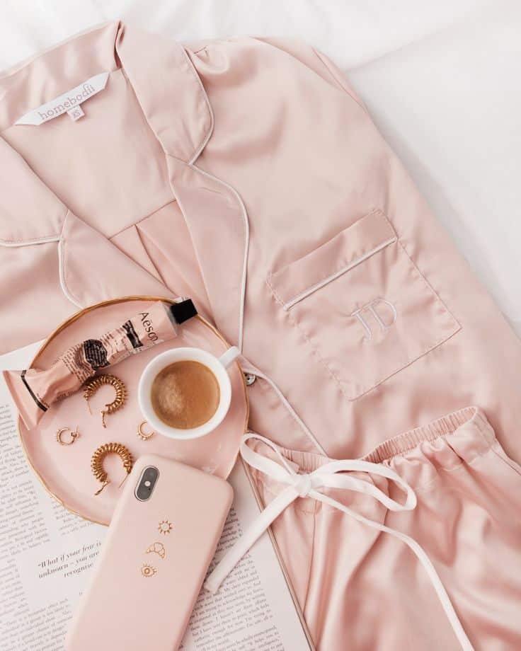 Soft pink silk pajamas, coffee, smartphone, hair accessories, and cosmetics on a cozy bed setting evoke relaxation and luxury.