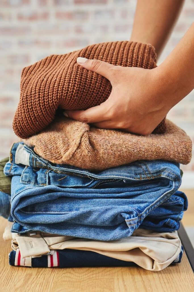 Person stacking folded sweaters and jeans on wooden table, displaying cozy wardrobe organization and casual fashion essentials.