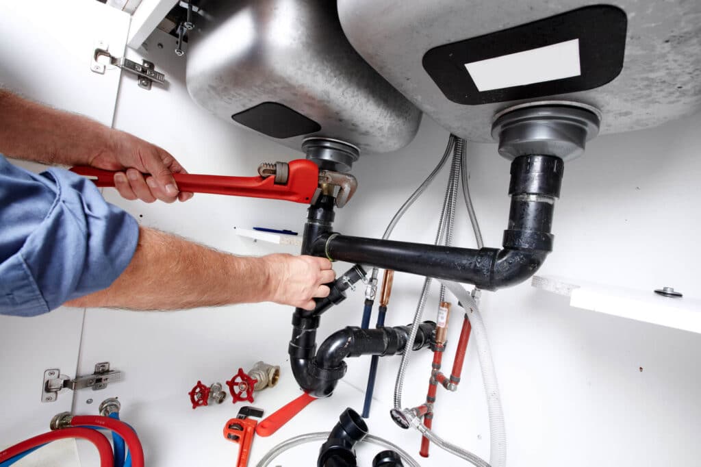 Plumber using a wrench to repair pipes under a kitchen sink, showing tools and fittings. Ideal for plumbing services or DIY repair articles.