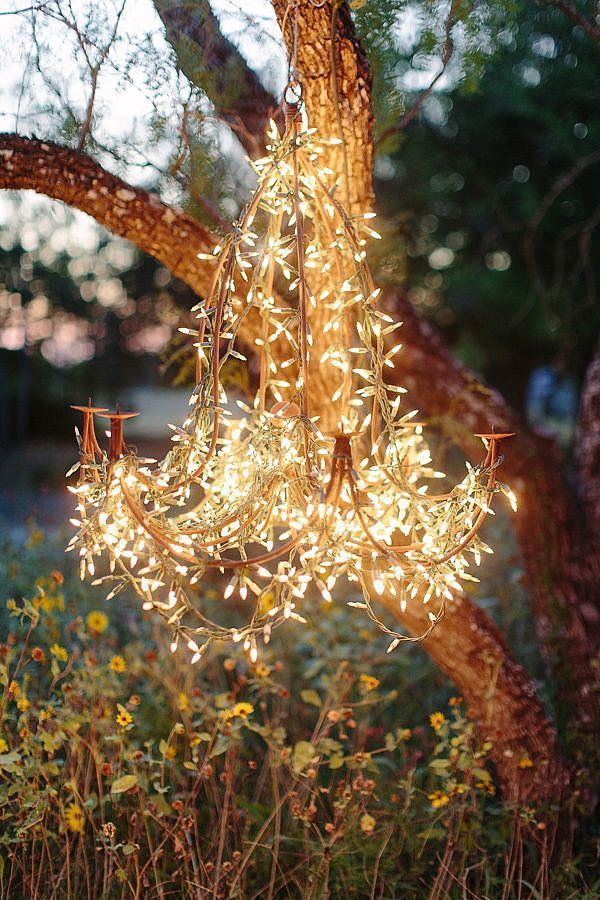 How great would it look if you wrapped lights around a chandelier for _ 28 Breathtaking Ways to Decorate With Christmas Tree Lights Rustic outdoor chandelier adorned with twinkling fairy lights hanging from a tree among wildflowers at dusk. | Sky Rye Design Rustic outdoor chandelier adorned with twinkling fairy lights hanging from a tree among wildflowers at dusk.