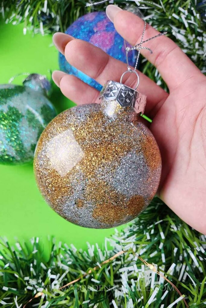 How to Make Glitter Ornaments With Regular Glue_ Hand holding a glittery gold and silver Christmas ornament against a green background with festive decorations. | Sky Rye Design Hand holding a glittery gold and silver Christmas ornament against a green background with festive decorations.