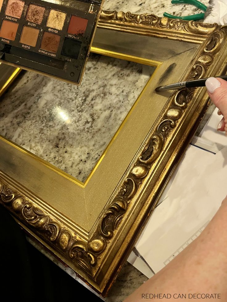 Hand applying black paint on ornate gold frame using eyeshadow palette for a DIY makeover project on a granite table.
