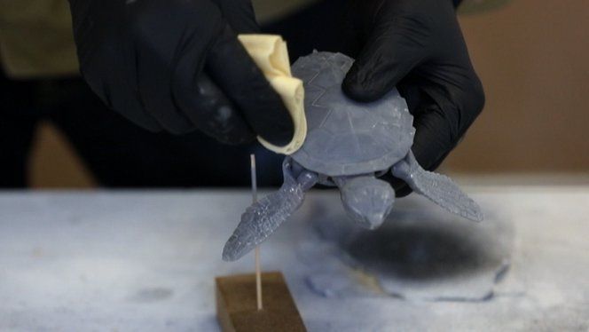 Person polishing a 3D-printed turtle model with gloves, focusing on detail work, on a workbench.