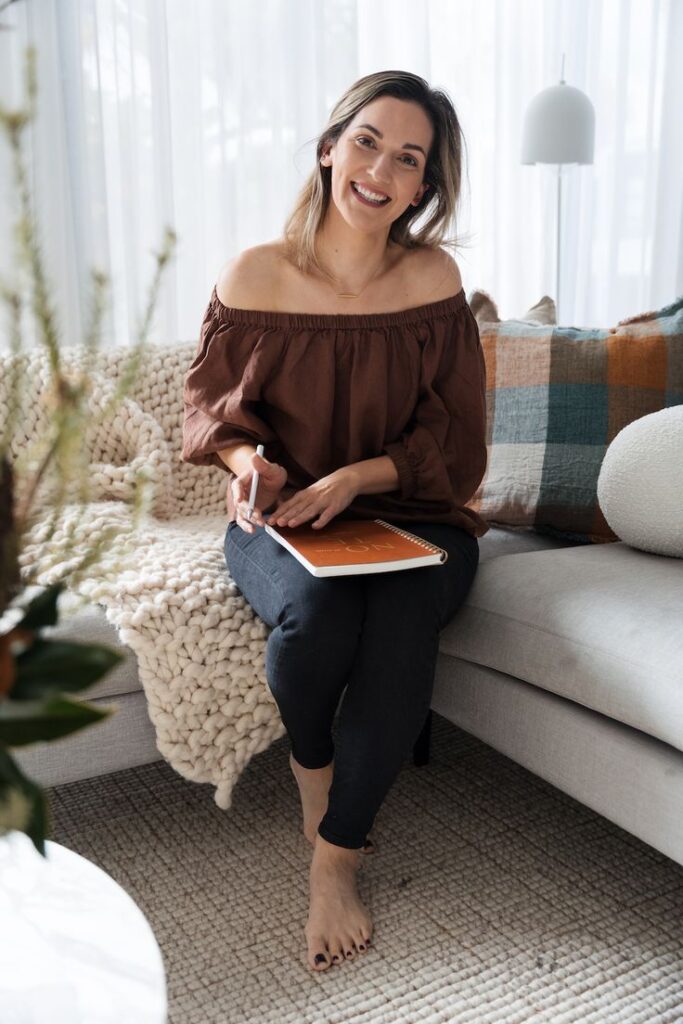 Woman sitting on a cozy couch holding a notebook and pen, smiling at the camera in a bright living room setting.