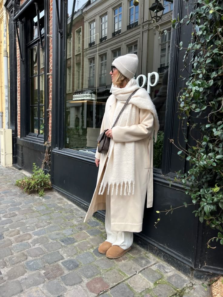 Woman in cozy winter outfit with a beige coat, scarf, and beanie, standing on a cobblestone street near a shop.