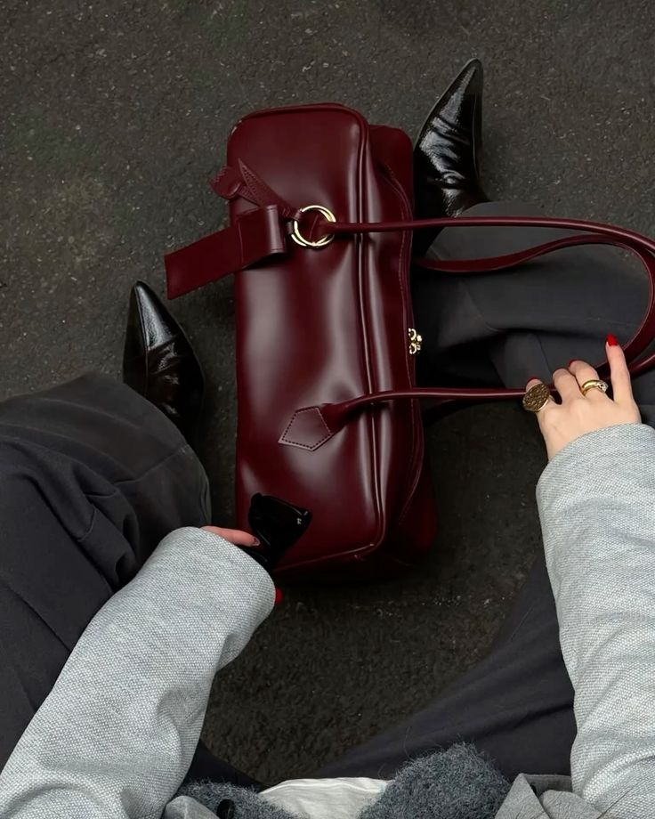 Person holding maroon leather handbag with stylish black boots visible, dressed in gray outfit and wearing a statement ring.