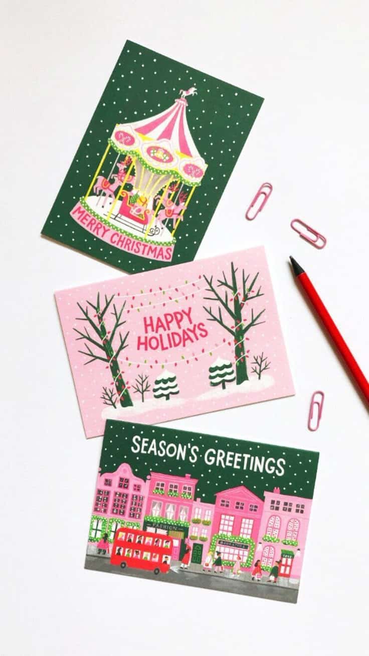 Colorful Christmas cards with festive designs and holiday messages, surrounded by a red pencil and pink paperclips on a white surface.