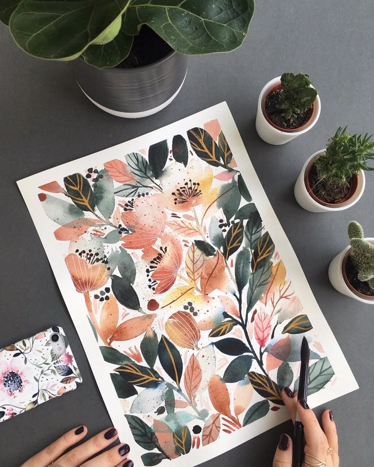 Inga Buive - Painting While My Son Naps LAURA HORN ART Colorful floral watercolor painting on a table surrounded by potted plants and a floral phone case. Hands holding a black paintbrush. | Sky Rye Design Colorful floral watercolor painting on a table surrounded by potted plants and a floral phone case. Hands holding a black paintbrush.