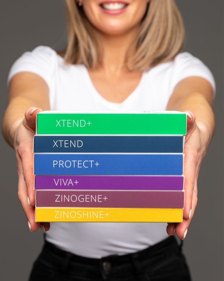 Inspire Change in Life _ Zinzino Woman holding stack of colorful health supplement boxes labeled XTEND+, PROTECT+, VIVA+, ZINOGENE+, ZINOSHINE+. | Sky Rye Design Woman holding stack of colorful health supplement boxes labeled XTEND+, PROTECT+, VIVA+, ZINOGENE+, ZINOSHINE+.