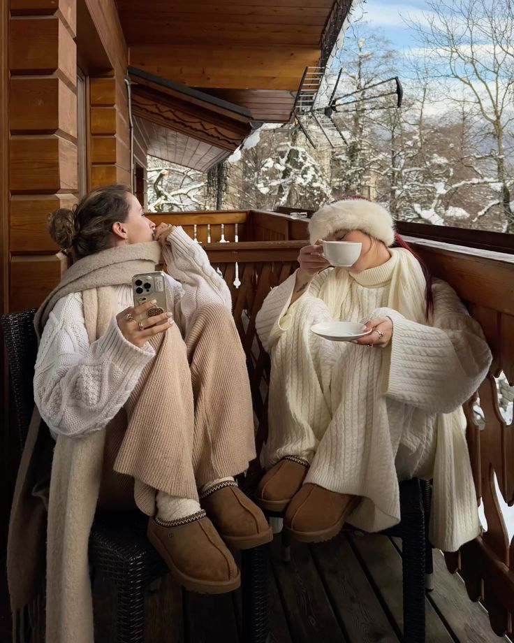 Two people in cozy winter outfits enjoy hot drinks on a snowy cabin balcony, surrounded by picturesque forest views.