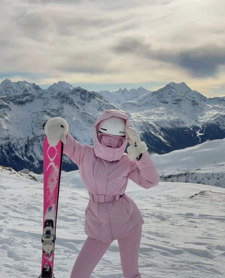 Skier in pink outfit poses on snowy mountain with skis, surrounded by breathtaking alpine scenery and overcast sky. Winter adventure.