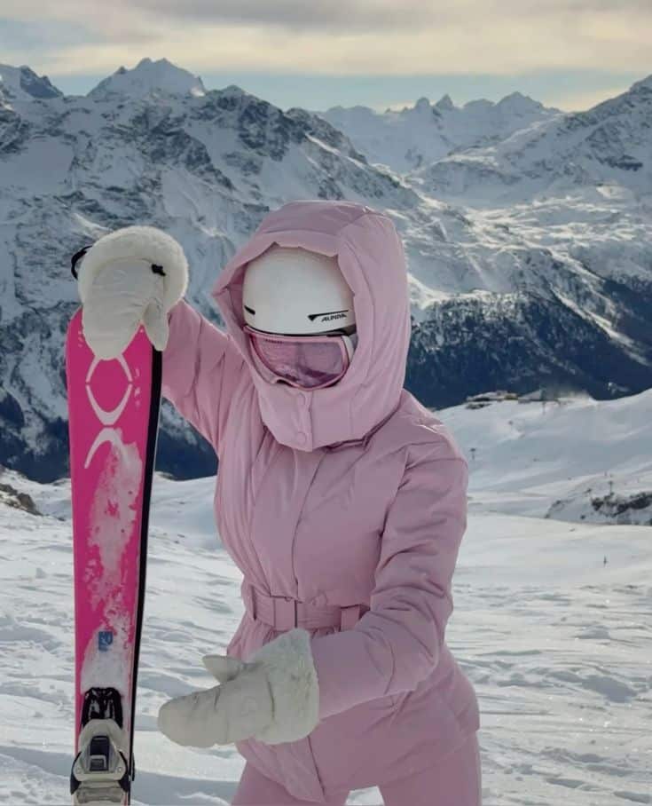 Person in pink ski outfit holding skis on snowy mountain with stunning alpine backdrop.