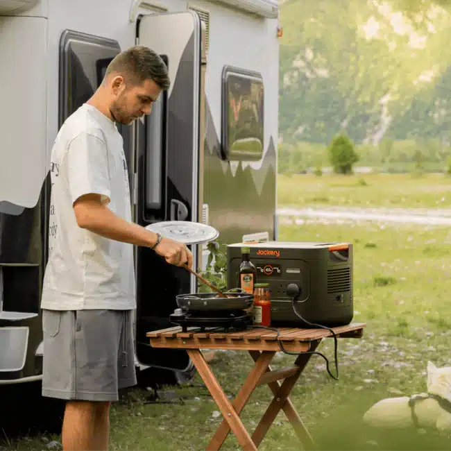 Jackery_Explorer_3000_v2_Portable_Power_Station_3 Man cooking outdoors with a portable power station next to a camper van in a scenic grassy area. | Sky Rye Design Man cooking outdoors with a portable power station next to a camper van in a scenic grassy area.