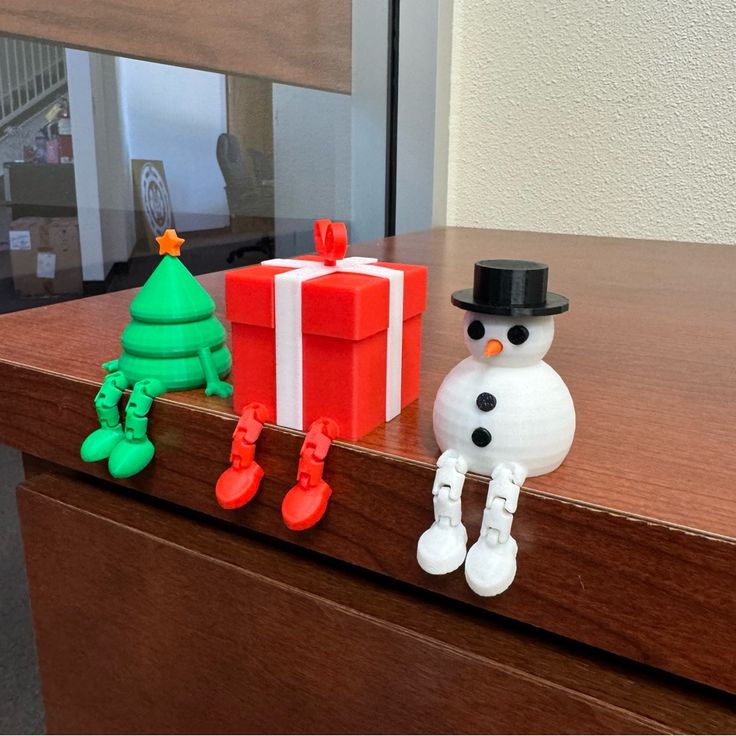 Adorable festive figures: a Christmas tree, gift box, and snowman with dangling legs sit playfully on a wooden cabinet edge.