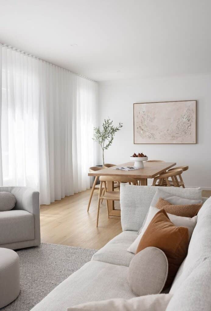 Modern living room with light wood dining set, white curtains, and neutral cushions for a minimalist aesthetic.
