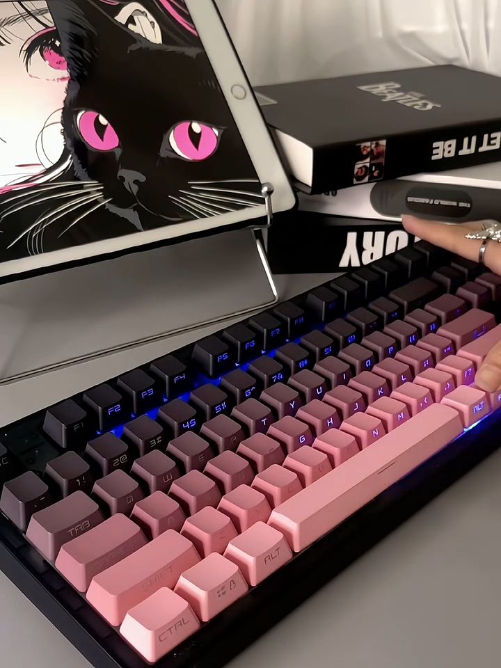 Pink and black mechanical keyboard with a digital tablet displaying anime art, alongside a stack of books and a hand adorned with rings.