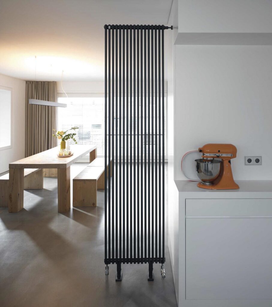 Modern kitchen with vertical black radiator, orange mixer on counter, and wooden dining set in bright, minimalist space.