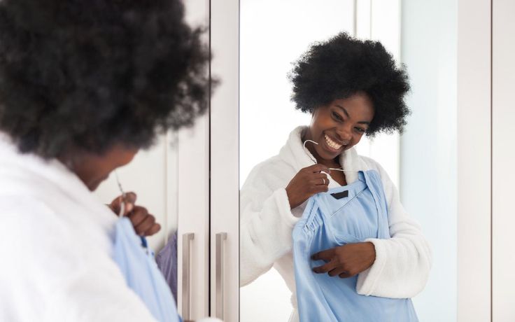 Woman smiling at a mirror while holding a blue dress on a hanger, reflecting joy and style in a modern dressing room.