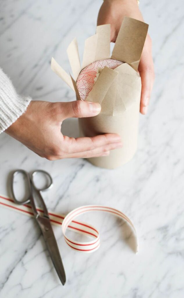Live Life Like You Mean It_ Person wrapping a cylindrical gift with paper on a marble surface, surrounded by scissors and striped ribbon. | Sky Rye Design Person wrapping a cylindrical gift with paper on a marble surface, surrounded by scissors and striped ribbon.