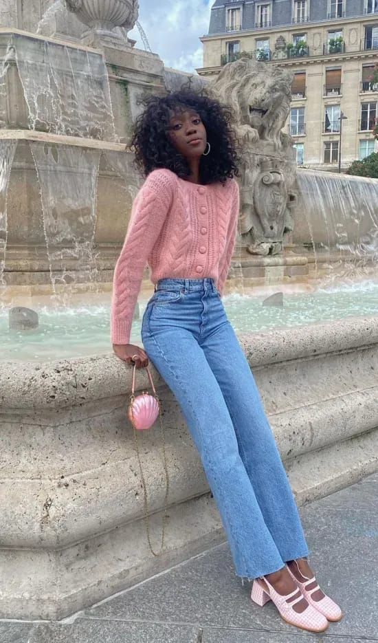 Stylish woman in pink sweater and jeans sits by a fountain holding a pink handbag, with historic architecture in the background.