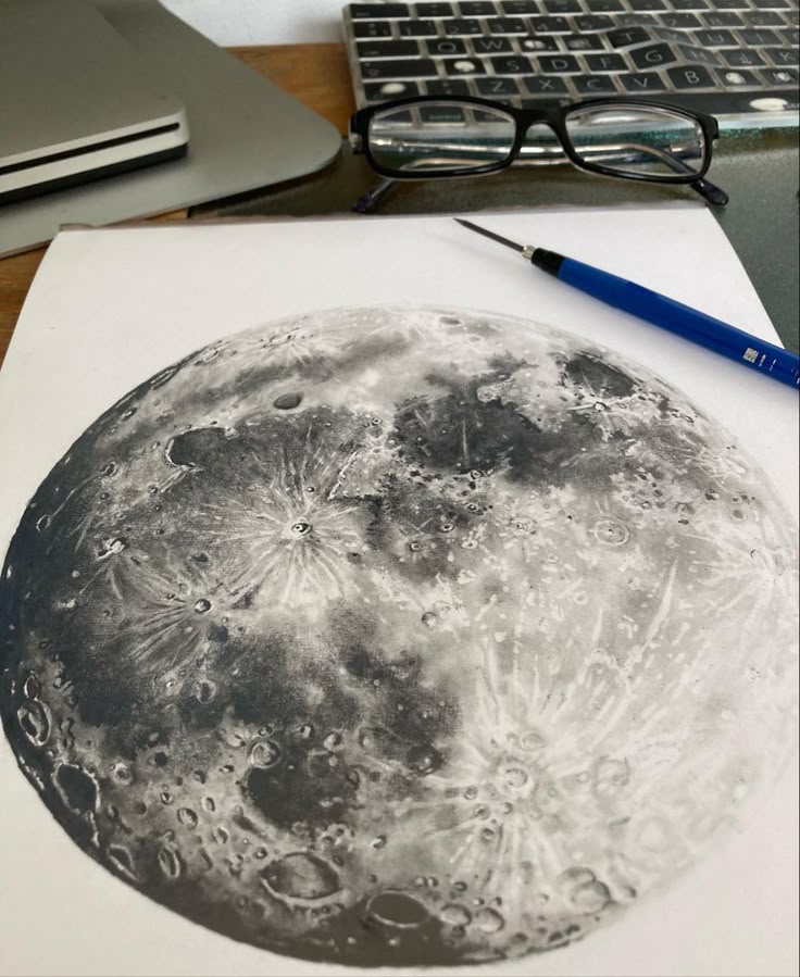 Detailed moon sketch on paper, surrounded by a pencil, glasses, and a laptop keyboard in a workspace setting.