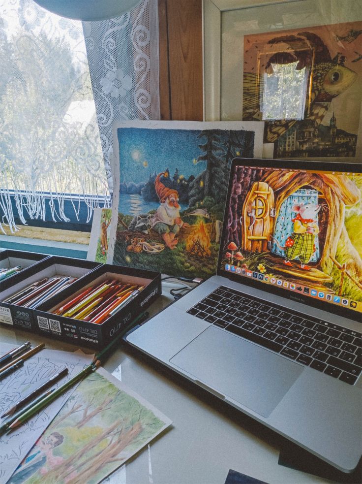 Artistic workspace with colored pencils, sketches, and vibrant illustrations on a laptop and canvas by a sunny window with lace curtains.