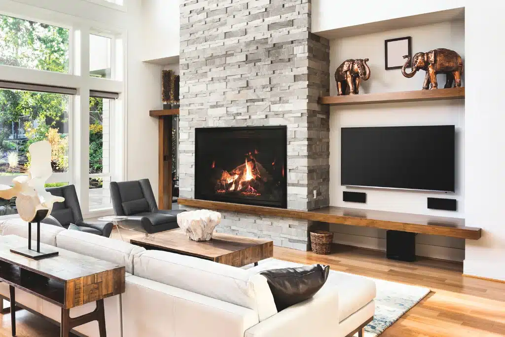 Modern living room with a stone fireplace, wall-mounted TV, and stylish decor. Ample light from large windows. Cozy seating area.