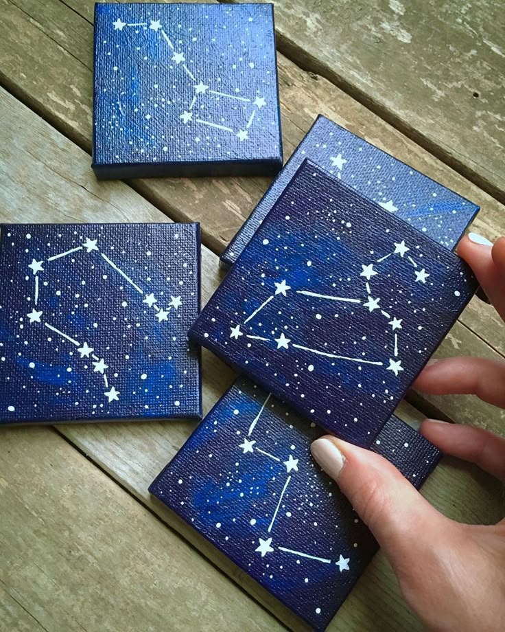Hand holding one of five small canvas paintings of constellations on wooden boards, showcasing a starry night theme.