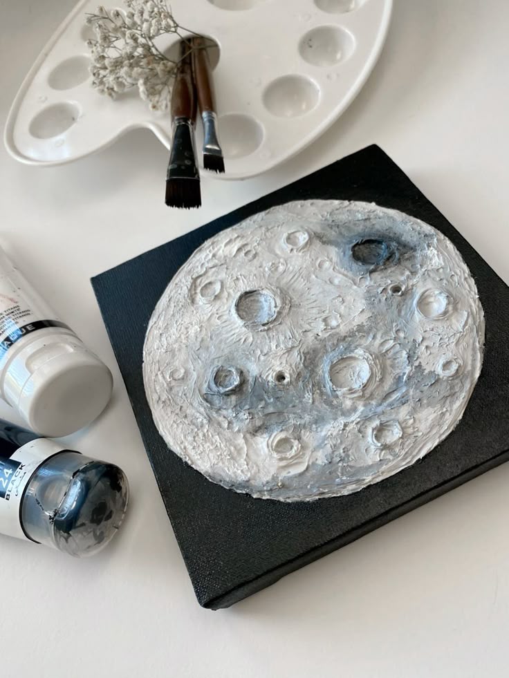 Textured moon painting on canvas beside paint supplies and brushes, showcasing detailed lunar craters and a creative artistic setup.