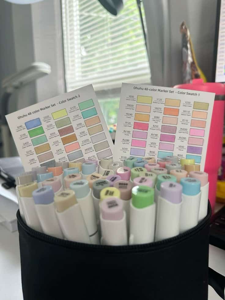 Ohuhu 48-color marker set with color swatches, in a black holder on a desk near a window, perfect for artists.