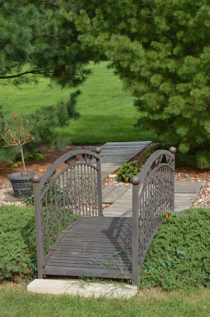 Decorative wooden bridge in a lush garden with greenery, surrounded by evergreens and a pebble pathway.
