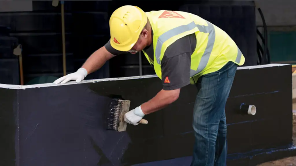 Worker applying waterproofing coat to concrete with a brush, wearing a safety vest and hard hat on a construction site.