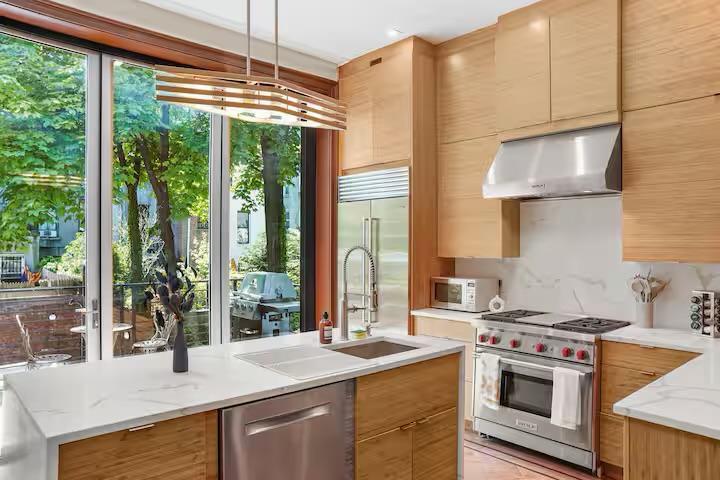 Modern kitchen with stainless steel appliances, wooden cabinets, and large windows overlooking a garden patio with a grill.